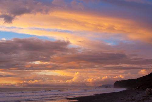 Spektakuläre Wolkenformation über den Wellen an einem steinigen Strand beleuchtet von einem rötlichen Sonnenuntergang